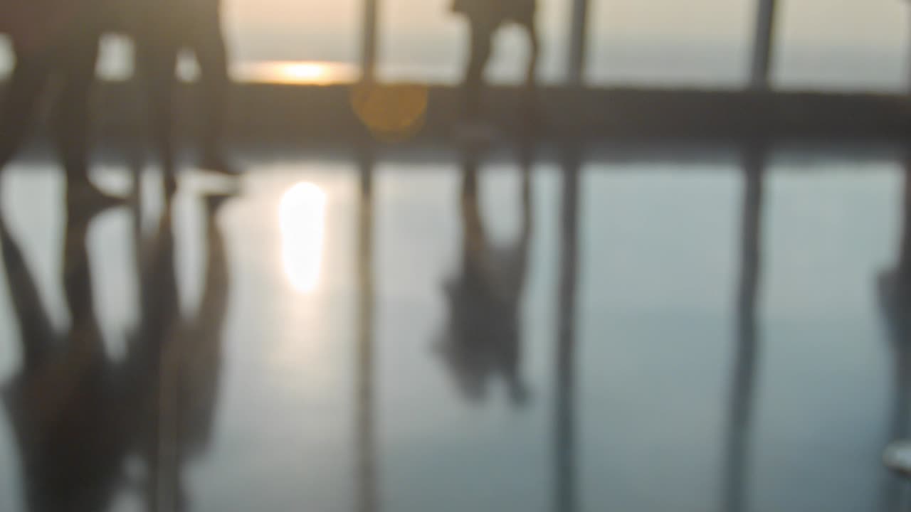 Persone irriconoscibili camminano su un pavimento riflettente all'interno di una stanza con grandi finestre e sole che entra direttamente, coppia che si abbraccia, ombre e piedi che si muovono - telecamera a mano.