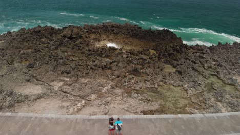 vista aérea de una pareja caucásica en la costa observando el hermoso paisaje marino azul tropical con olas chocando contra las rocas en bali