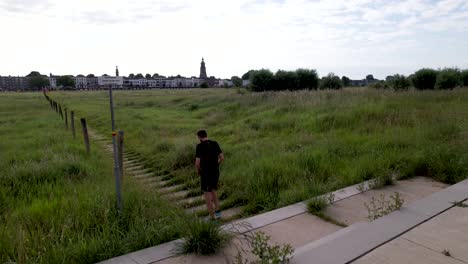 Start-and-aerial-following-of-male-trail-runner-Dutch-river-IJssel-floodplains-with-countenance-of-tower-town-Zutphen