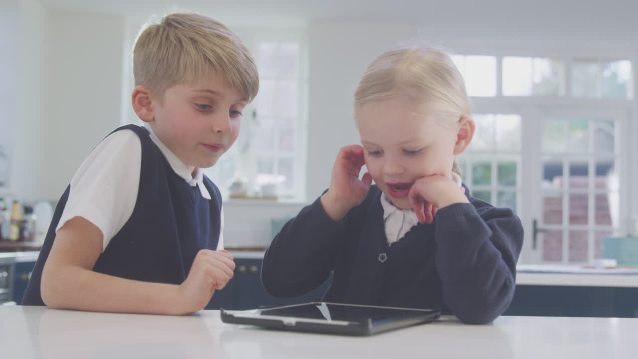 dos niños vestidos con uniforme escolar en la cocina jugando con una tableta digital en el mostrador