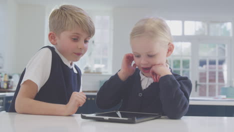 Dos-Niños-Vestidos-Con-Uniforme-Escolar-En-La-Cocina-Jugando-Con-Una-Tableta-Digital-En-El-Mostrador.