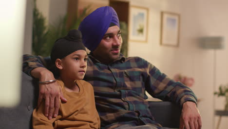 Padre-Sikh-Y-Su-Hijo-Pequeño-Con-Turbantes-Sentados-En-El-Sofá-En-Casa-Hablando-Juntos