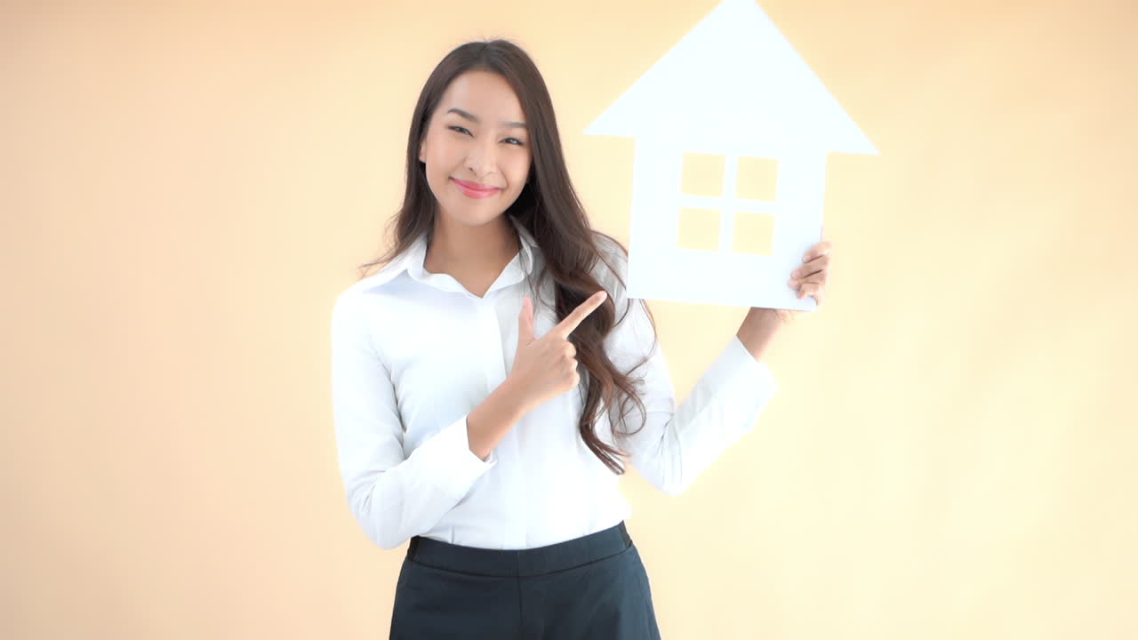 A pretty young woman holds up a cutout house then points to it