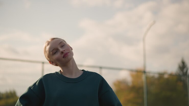 Young lady in green sweater with hand on waist, working out by turning head from left to right, sunlight creates golden glow around her