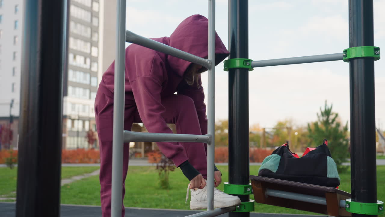 Jogger confidently ties shoelace on iron bar corner after placing gym bag then adjusts hoodie before beginning vigorous outdoor fitness session in urban park under clear sky near modern building
