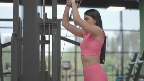 a hispanic brunette woman in a pink suit pulls a rope in a crossover with her hands to train her shoulders. shoulder workout in a trainer. professional woman instructor