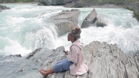black lady on her phone sitting on the rocks near the edge of a waterfall (natural bridge lower falls) and beautiful scenic blue river in yoho national park