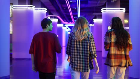 group of three friends walking by cinema hall with neon interior, rear view