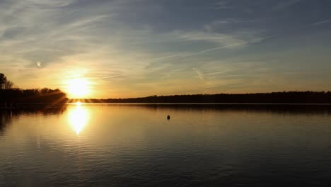 Peaceful-and-quiet-lake-with-smooth-water-surface-reflects-golden-glow-of-setting-sun-and-light-thin-clouds-in-the-sky