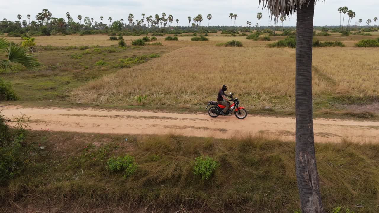 Un drone orbita a destra mentre segue un motociclista su una strada sterrata nella Cambogia rurale