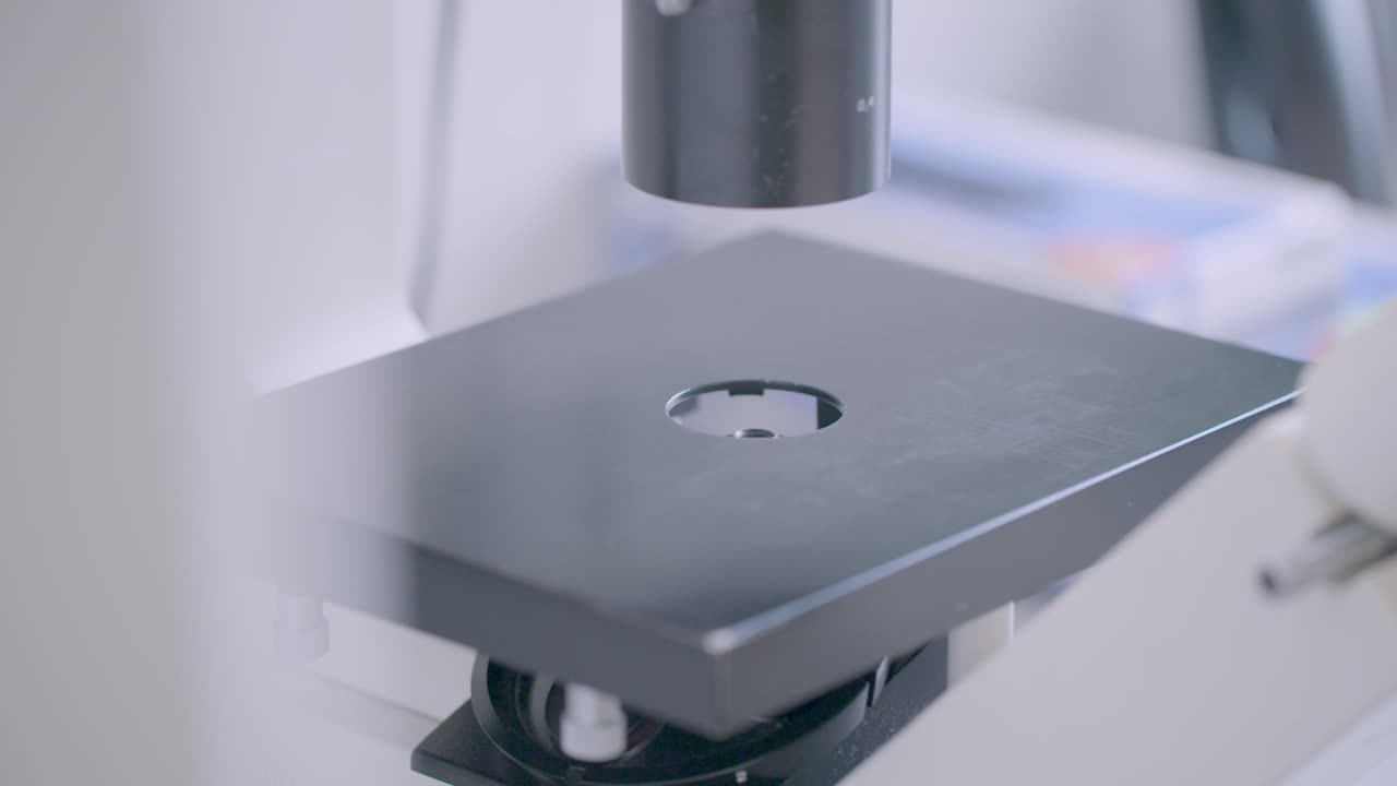 Close-up of a gloved hand positioning a petri dish with red media beneath a microscope objective in a modern lab.