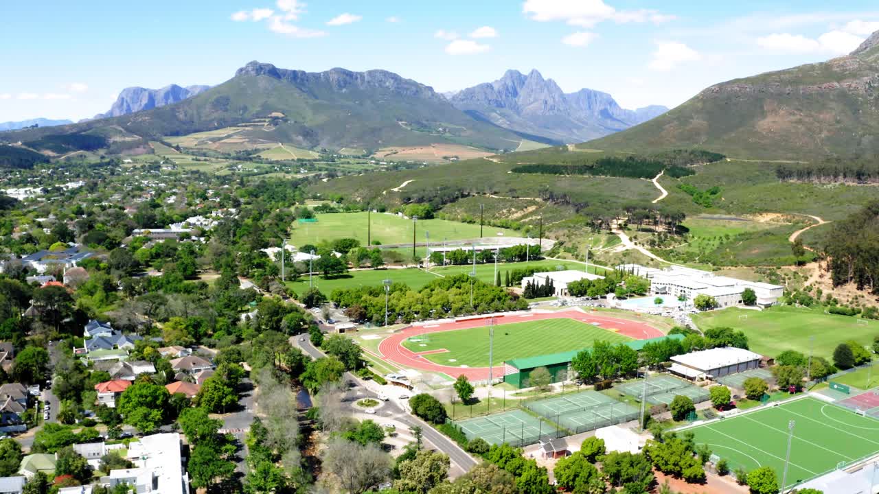 rotación de drones aéreos, estadio de atletismo deportivo de la universidad universitaria, pista y campo, canchas de tenis, montañas, árboles y vecindario en el fondo, stellenbosch, coetzenburg