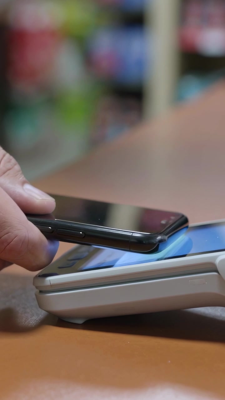 Video verticale: Una mano che tiene uno smartphone tocca un terminale POS alla cassa di un negozio, effettuando un pagamento.