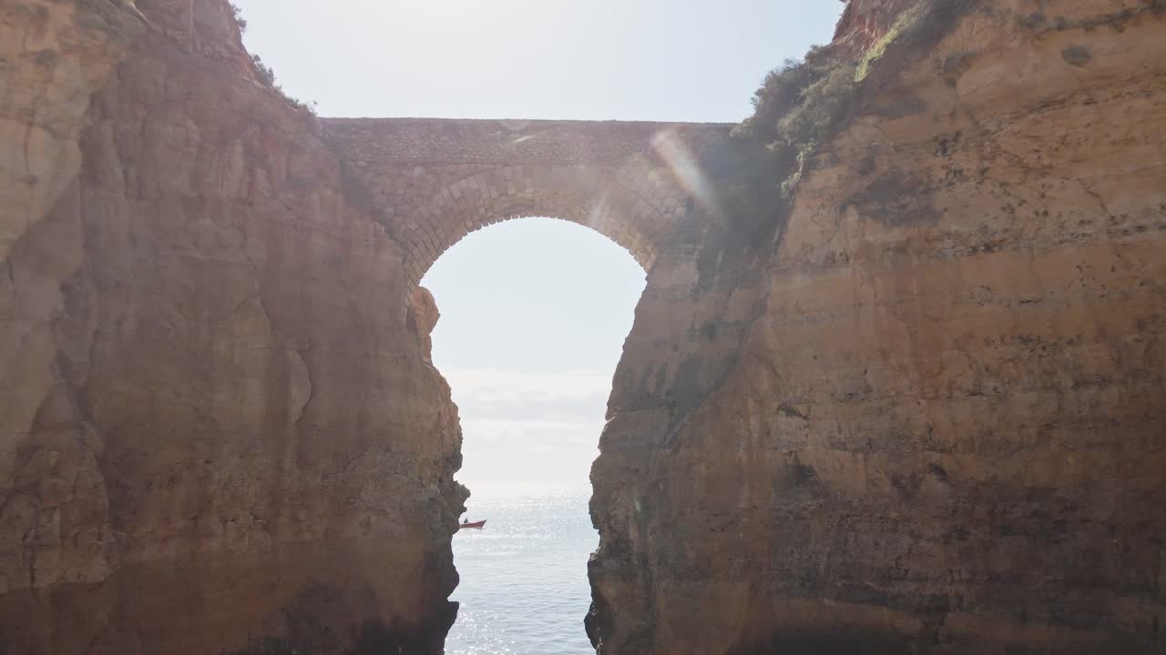 Aerial drone flies forward, passing through a dramatic natural stone archway between towering limestone cliffs, revealing the sparkling sea beyond near Lagos, Portugal