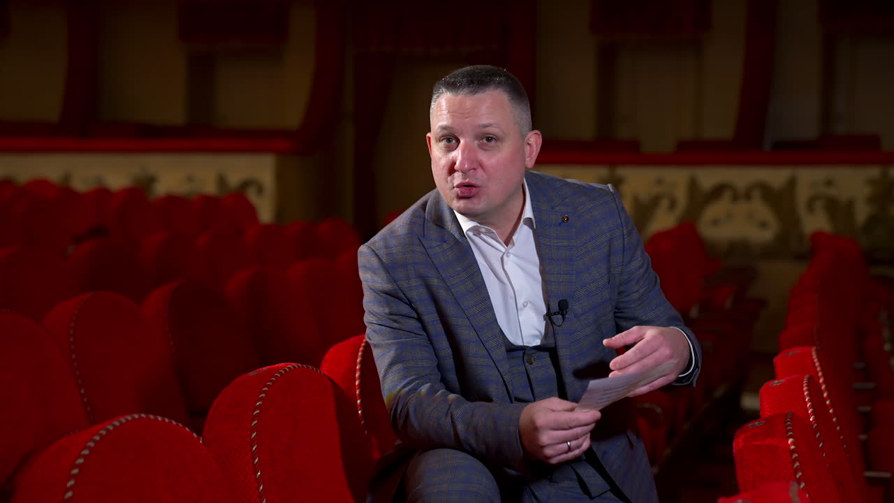 Performer announces future theater season. Actor is sitting in empty theater on red velvet chair with sheet of paper in hands.