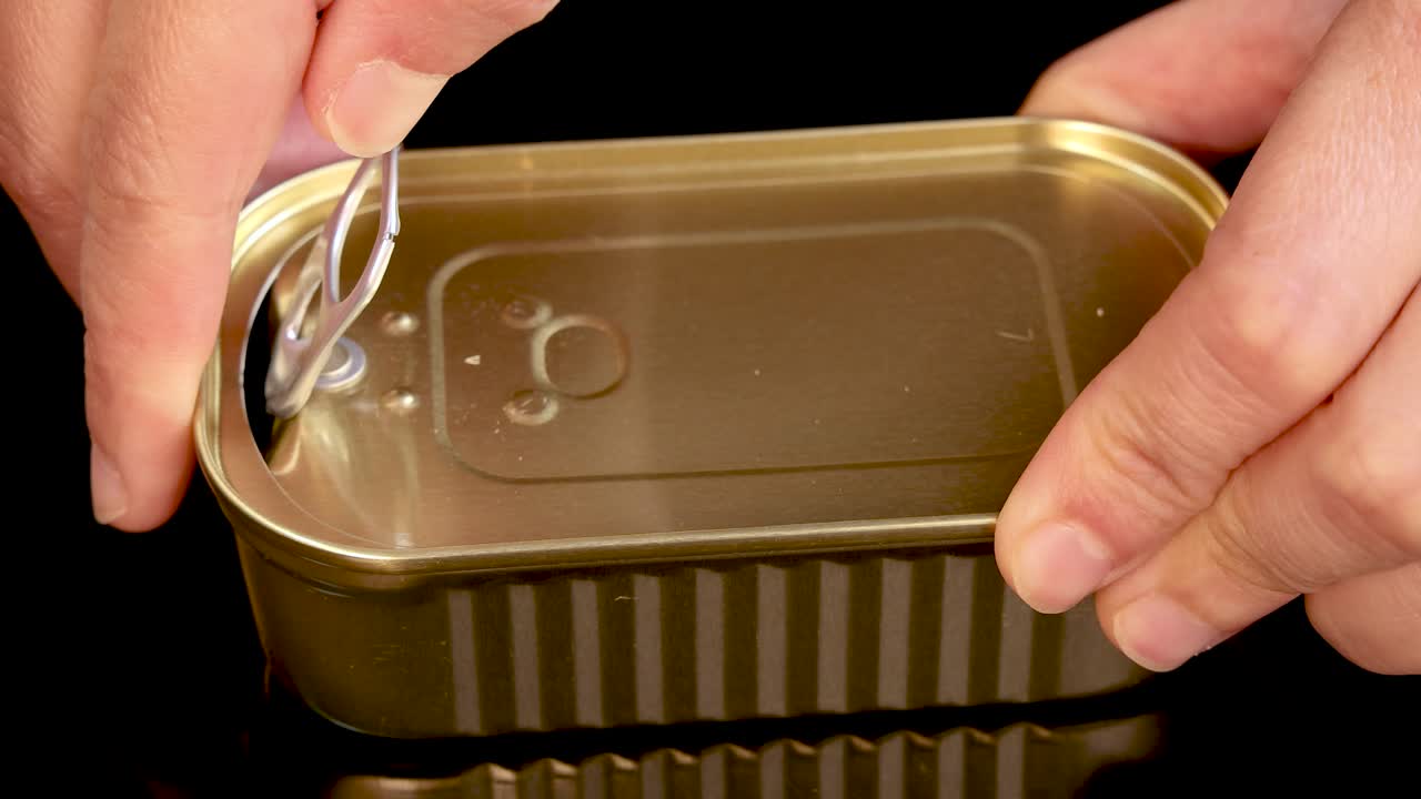 A close-up sequence shows hands opening a rectangular sardine can with a pull tab, under bright lighting on a black background, revealing the metal lid peeling back