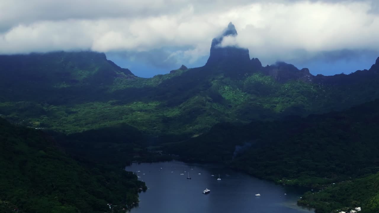 Dramatiska berget Rotui ovanför Opunohu-bukten i Moorea, Franska Polynesien, flygdrönarvy med segelbåtar som vilar i bukten nedanför branta gröna sluttningar och låga moln cirkulerar åt höger.
