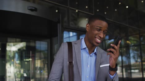 Smiling-african-american-businessman-using-smartphone-outside-of-modern-office-building