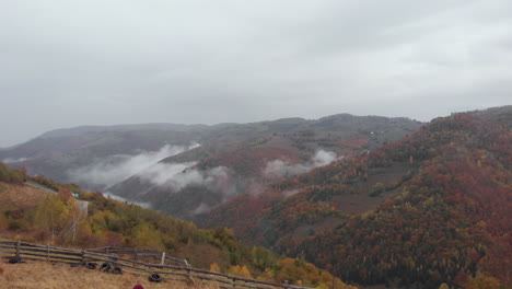 A-couple-of-hikers-holding-hands-walking-on-top-of-a-countryside-hill-sightseeing-autumnal-colors-valley-covered-with-tree-forest,-drone-flying-forward