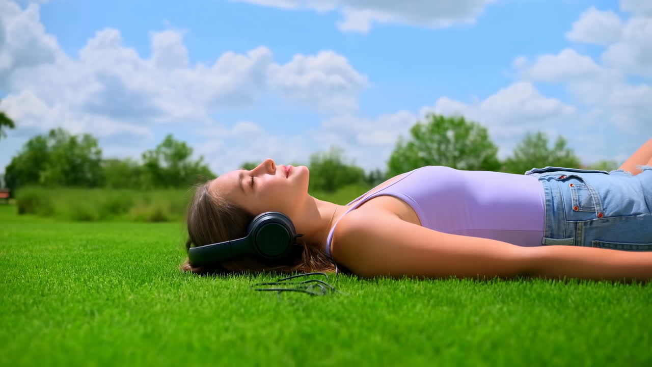 Junge weiße Frau liegt auf Gras und hört Musik mit schwarzen Kopfhörern, trägt ein lavendelfarbenes Tanktop