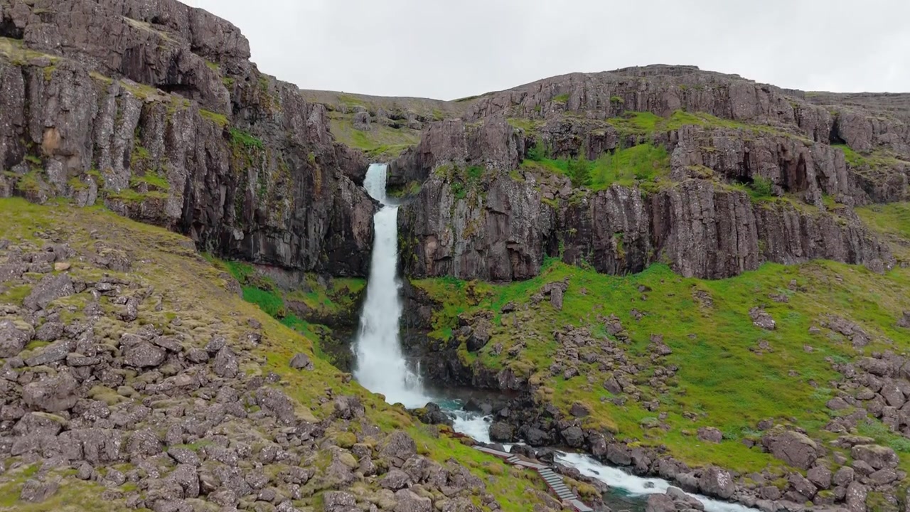 Droneoptagelse med opadgående tilt over Snædalsfoss vandfald og klippefyldt kløft i det østislandske landskab.