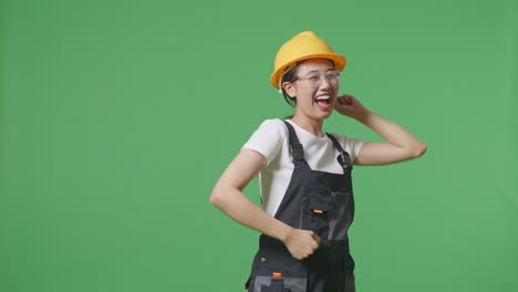 asian woman worker wearing goggles and safety helmet singing and dancing while standing in the green screen background studio