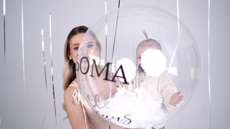 happy smiling toddler baby in mom's hands holding a big transparent balloon. mother and child footage in studio.