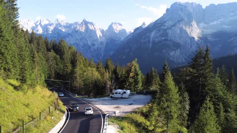 Eine-Drohnenaufnahme-Einer-Bergstraße-In-Den-Dolomiten-Mit-Vorbeifahrenden-Autos-Und-Einem-Am-Straßenrand-Geparkten-Wohnmobil-Inmitten-Eines-Von-Bergen-Umgebenen-Pinienwaldes-In-Norditalien