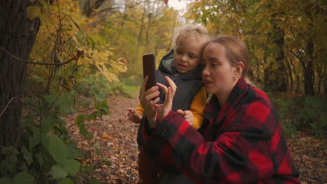 La-Madre-Y-El-Bebé-Caucásicos-Descansan-En-El-Bosque-Y-Ven-Fotos-En-Teléfonos-Inteligentes,-Fotografías-Móviles-De-La-Naturaleza-Descansando-Y-Aficiones-Los-Fines-De-Semana