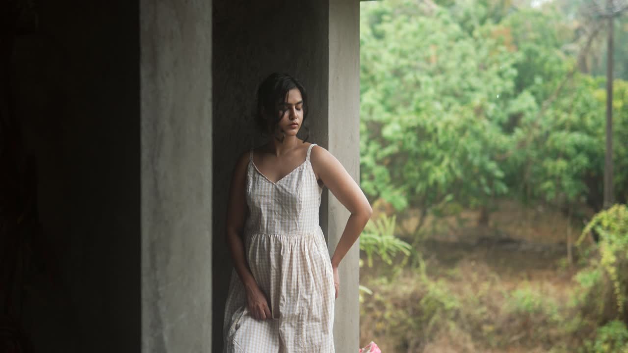 가운 드레스 를 입은 여자 가 농촌 의 문 앞 에 사려 깊게 서 있으며, 배경 에 푸른 초록색 이 있다