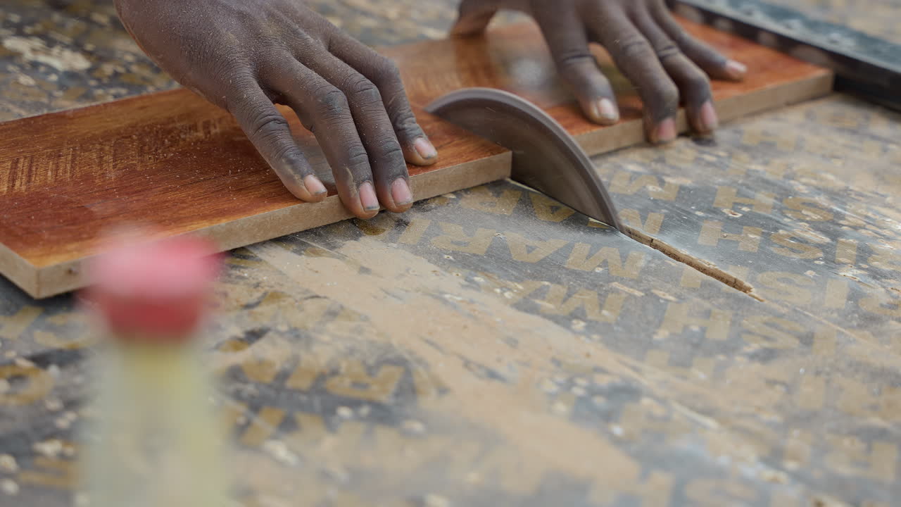 Black hands guiding hardwood across saw, apprentice carpenter trims plank with steady grip, blade slicing through grain, sawdust scattering on workbench, gritty workshop light, focused carpentry craft
