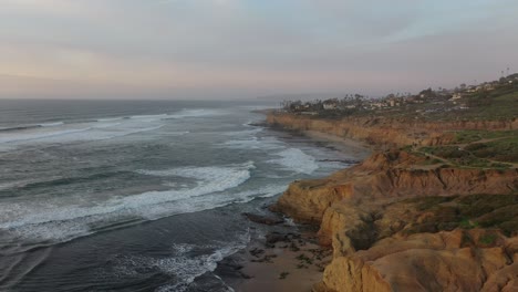 una impresionante toma de dron volando sobre los acantilados con las olas rompiendo debajo, capturando el poder puro de la naturaleza y la calidez del sol poniente
