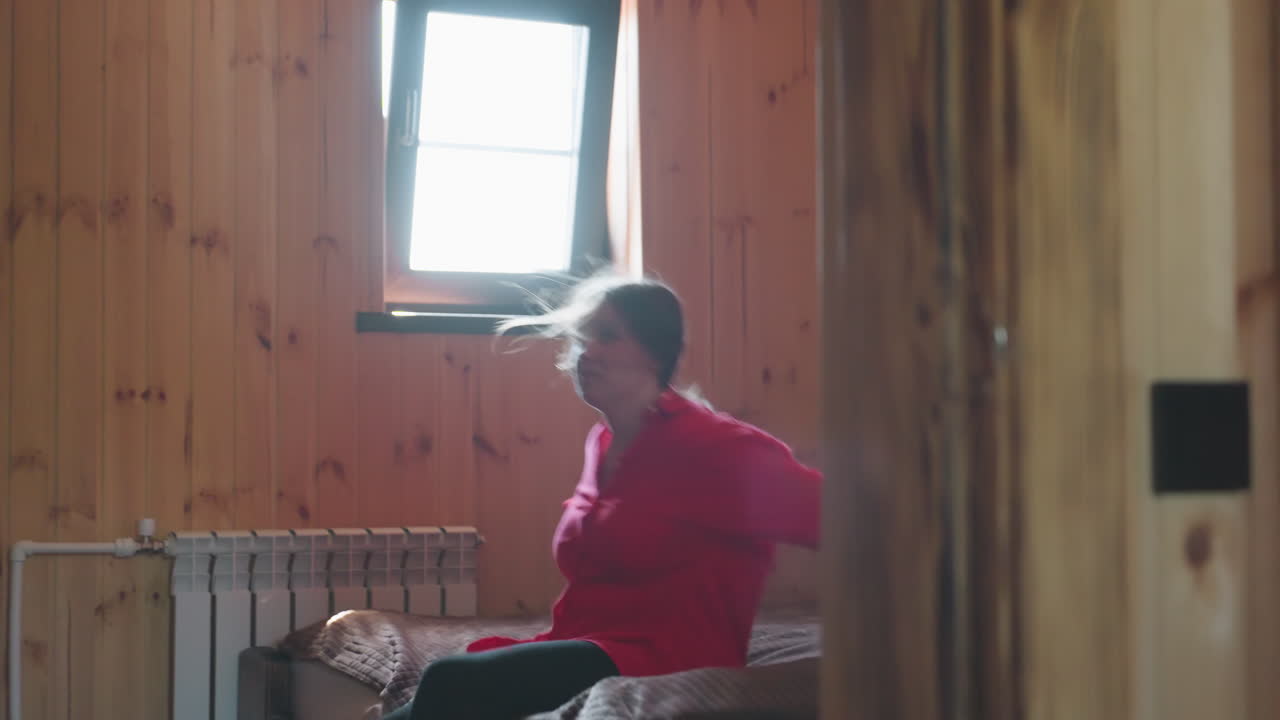joyful girl in red shirt jumps playfully onto bed inside cozy wooden room with bright daylight streaming through window, creating warm and carefree indoor atmosphere