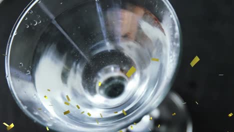 golden confetti falling over close up of olives falling into cocktail glass against black background