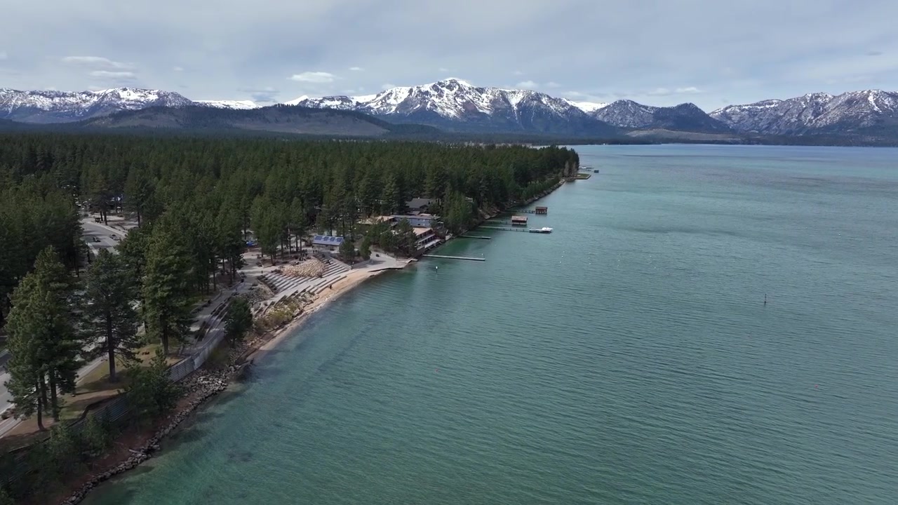 Auto che guidano lungo una strada costiera accanto ad acqua limpida e blu con una costa boscosa e montagne innevate a South Lake Tahoe, California, che mostrano un paesaggio alpino panoramico degli Stati Uniti. Sorvolo aereo.