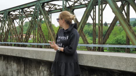 chica con gafas y suéter negro comprobando el teléfono con puente de tren de hierro en el fondo