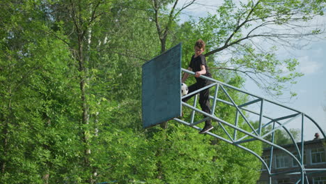 a young boy is seated on a blue iron outdoor structure, focusing intently as he stretches himself and looks downward, a worn soccer ball is stuck in the iron framework