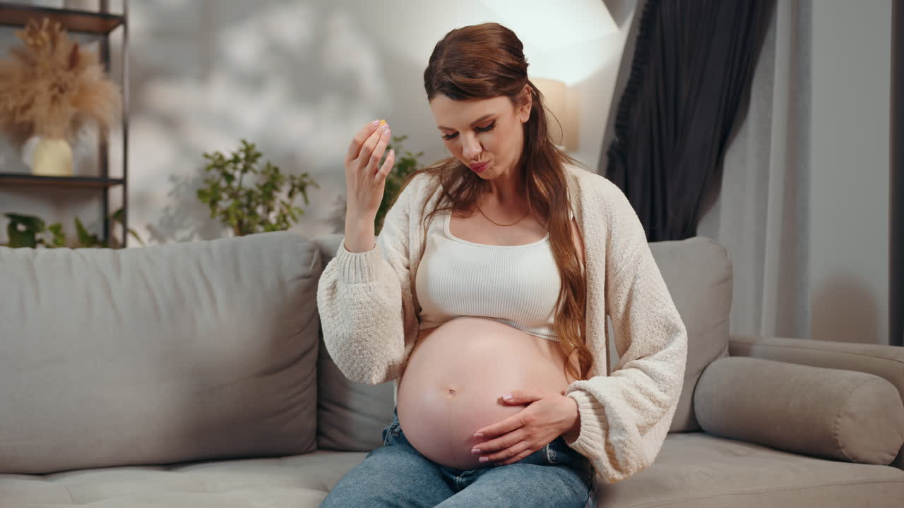 Pregnant woman sits on sofa holding belly and eating small snack feeling content and relaxed in cozy