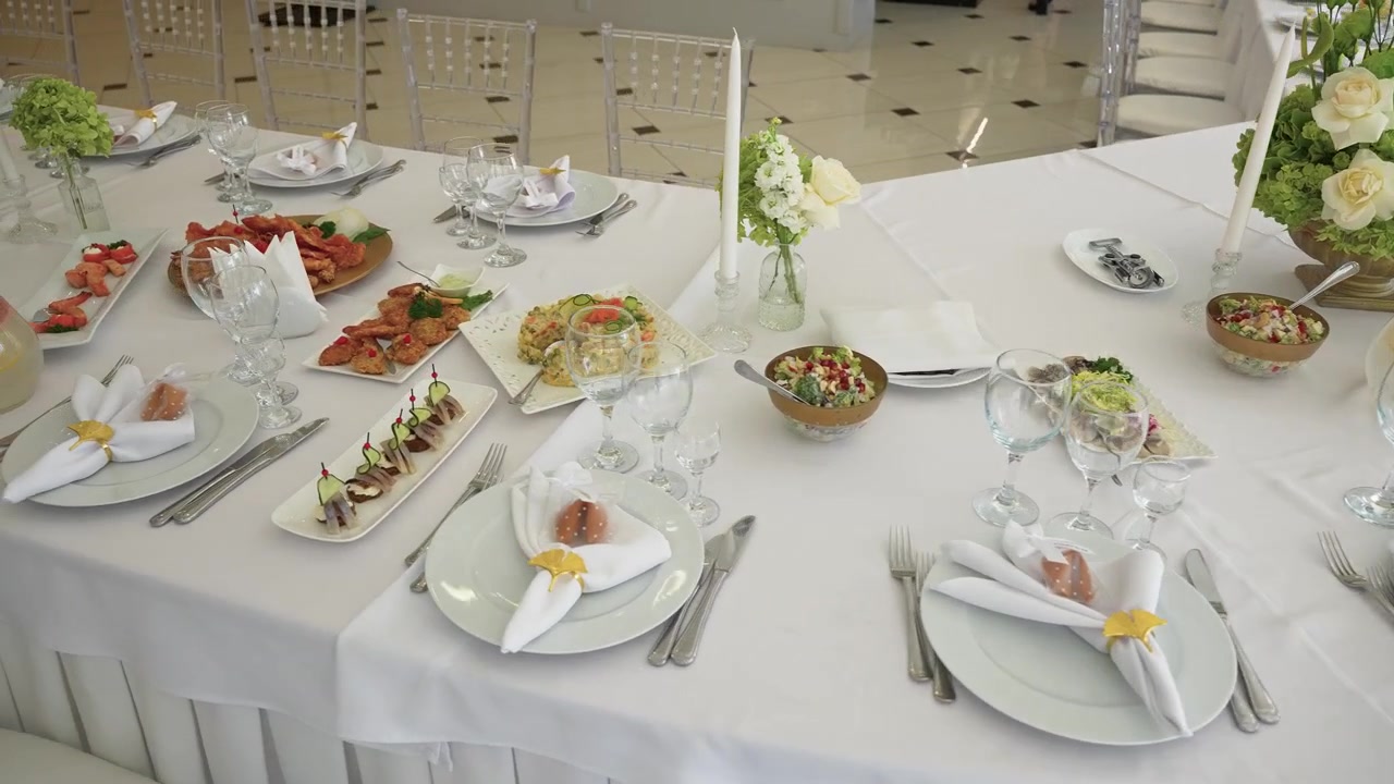 Weelderige witte feesttafel elegant gedekt met gastronomische hapjes, kristallen glaswerk, witte bloemen en kaarsen onder heldere, natuurlijke binnenverlichting. Statisch shot benadrukt fijne details