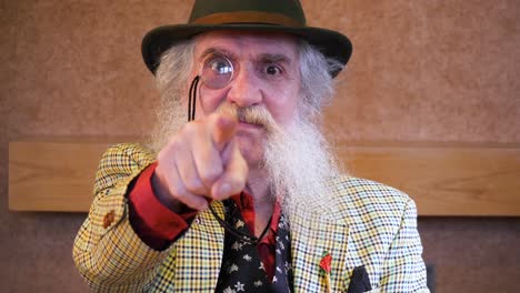 older man with beard, monocle and hat points at camera