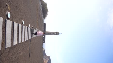 Mujer-Caminando-Sobre-Cenizas-Volcánicas-Con-El-Faro-De-Ponta-Dos-Capelinhos-Al-Fondo,-Faial,-Azores