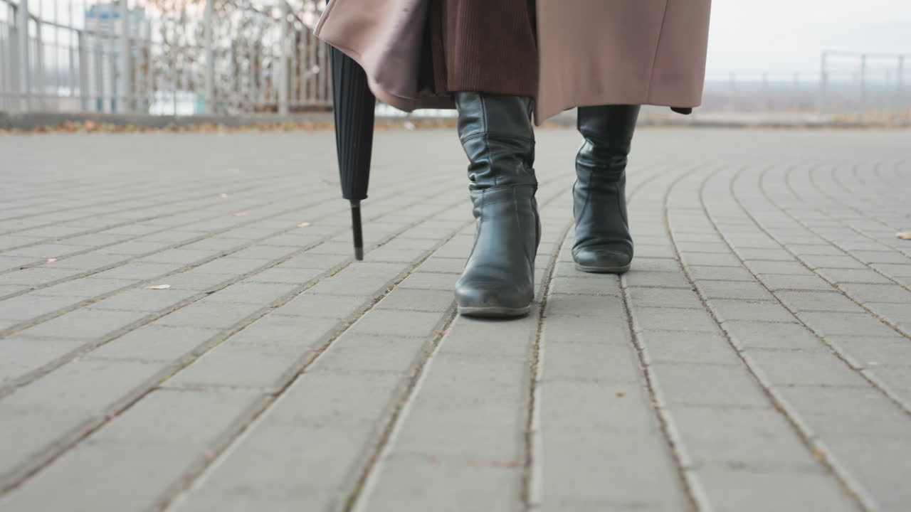 Sakin bir şehir parkında karo döşeli kaldırımda yürüyen, işe gidip gelen bir kadının botları ve şemsiyesinin yakın çekimi. Deri botları ve uzun paltosuyla yavaş, ölçülü adımlar atıyor, şemsiyesinin ucu yere sürtünüyor. Hava bulutlu.
