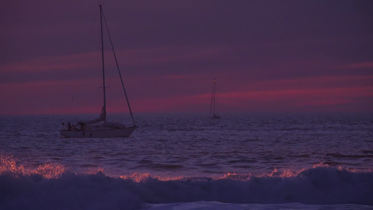 un velero pasa al atardecer por una playa del sur de california