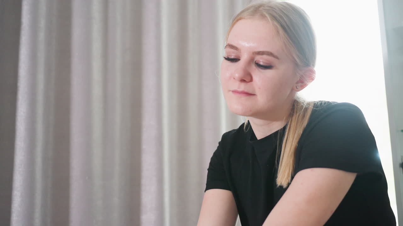 Close up of young female massage therapist in black shirt with soft smile and concentrated expression, working in bright wellness room with curtain background during relaxing therapy session