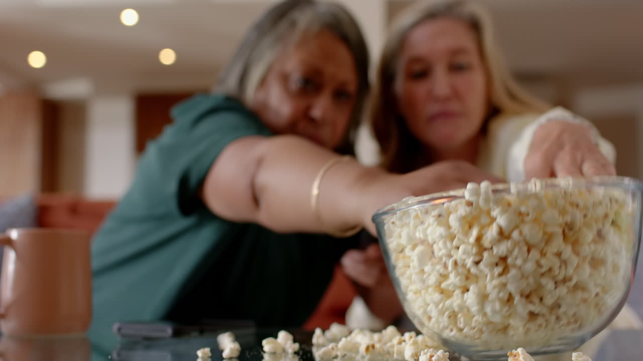 Eating popcorn, multiracial senior female friends enjoying time in cozy living room, at home