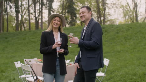 middle aged man and woman laughing and looking at camera while holding wine glasses in the park