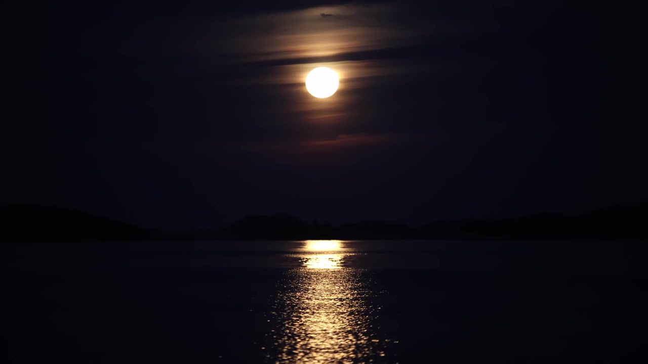 Notte buia con luna piena giallastra dietro nuvole sottili, luce scintillante che si riflette sulla superficie calma dell'oceano con leggere increspature.