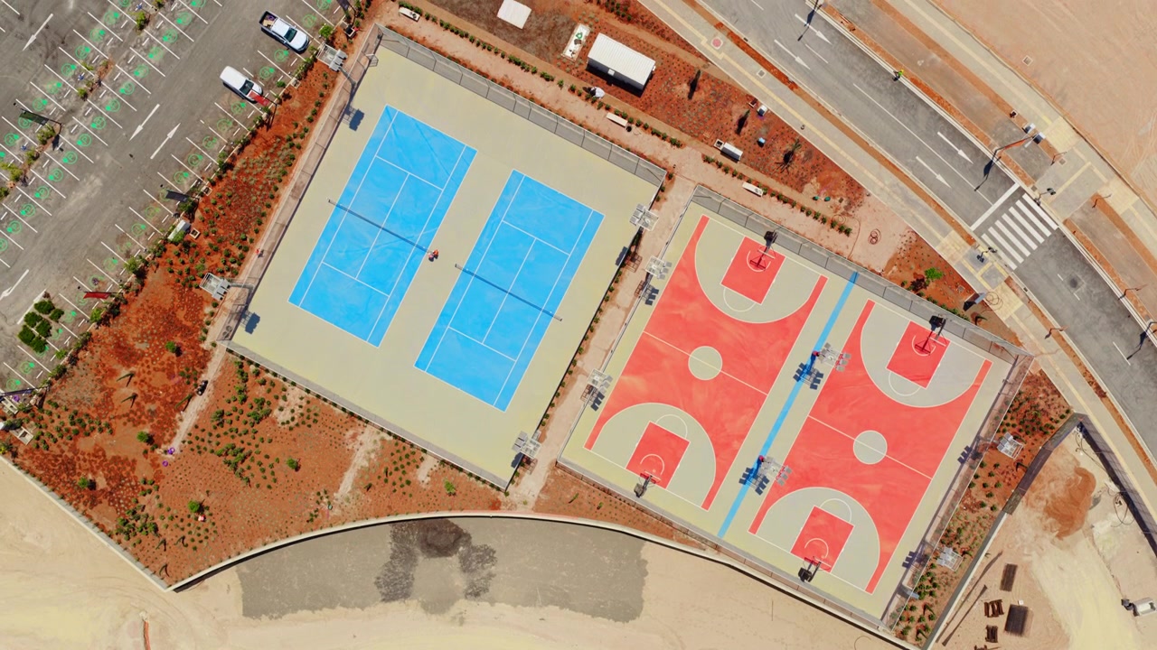 Vista aérea superior mostrando o progresso da construção de um parque público com quadras de basquete e tênis, desde a terraplanagem inicial até a área recreativa concluída e o estacionamento