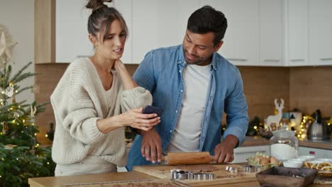 una pareja multiétnica haciendo panes de jengibre y usando teléfono móvil en la época de navidad.