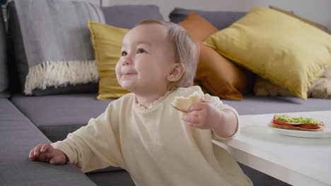 linda niña comiendo pan mientras está de pie junto al sofá en la sala de estar